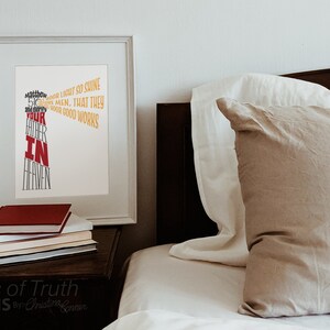 May include: A white framed print with a typography design that reads "Let your light so shine before men, that they may see your good works and glorify your Father in Heaven". The print is on a wooden nightstand next to a bed with a beige pillow and white bedding.