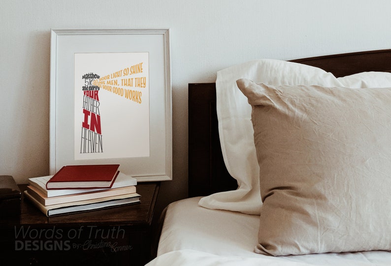 May include: A white framed print with a typography design that reads "Let your light so shine before men, that they may see your good works and glorify your Father in Heaven". The print is on a wooden nightstand next to a bed with a beige pillow and white bedding.