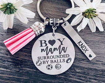 May include: A silver keychain with a round pendant that reads "Boy Mama Surrounded by Balls" and a rectangular tag with the name "Knox". The keychain has a pink tassel and a silver ring.