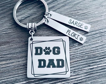 May include: Silver keychain with a square tag that says "DOG DAD" with a paw print in the middle. Two smaller tags are attached to the keychain, one says "SARGE" and the other says "FLOKI".