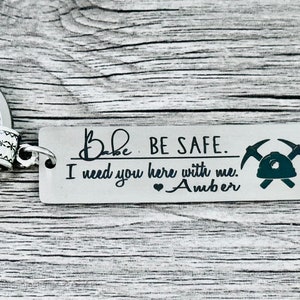 May include: Silver keychain with a black inscription that reads "Babe BE SAFE. I need you here with me. Amber" with a heart symbol and a black silhouette of a miner's helmet and pickaxe.
