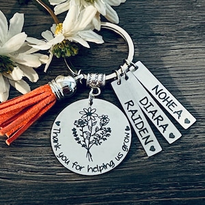 May include: Silver keychain with a round charm featuring a floral design and the text "Thank you for helping us grow".  The keychain also includes three rectangular tags with the names "Nohea", "Diara", and "Raiden" engraved on them.
