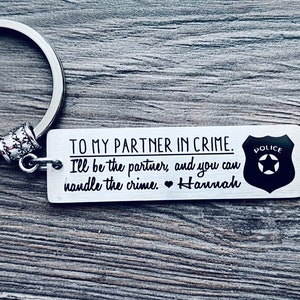 May include: A silver keychain with a black and white inscription that reads "To my partner in crime. I'll be the partner, and you can handle the crime. Hannah" and a black and white police badge with a star.