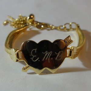 May include: A gold-toned bracelet with a double heart charm. The charm has an arrow through it and the initials "E.M.B." engraved on it.