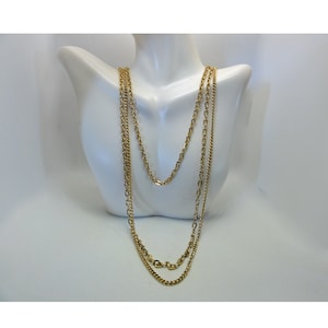 May include: A gold-colored necklace with three strands of varying chain styles. The necklace is displayed on a white mannequin bust. The chains have different link designs, creating a layered effect. The necklace is a classic jewelry piece.