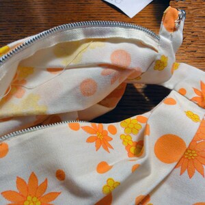 May include: A white fabric with a zipper closure and a floral pattern in shades of orange and yellow. The fabric has a textured surface and a subtle sheen.