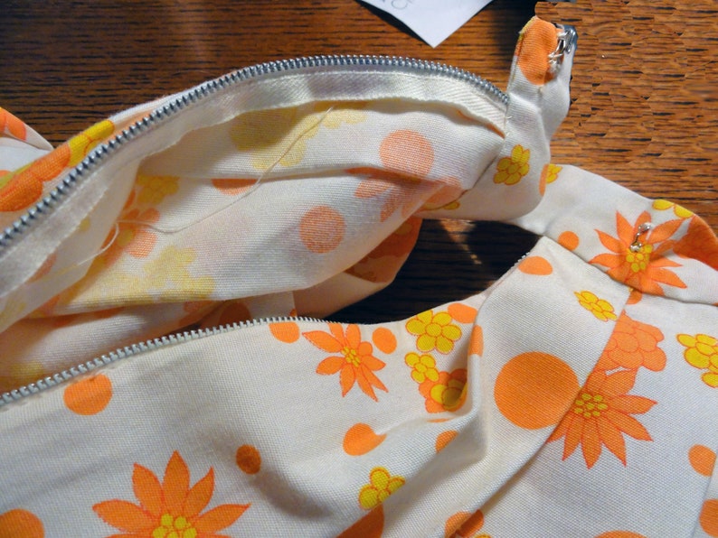 May include: A white fabric with a zipper closure and a floral pattern in shades of orange and yellow. The fabric has a textured surface and a subtle sheen.
