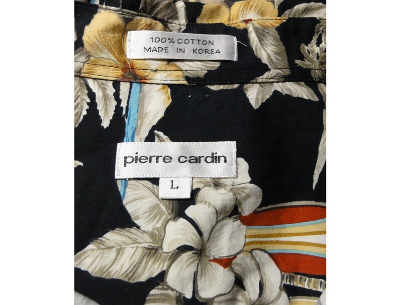 May include: A black button-up shirt with a floral print. The shirt has a white label that says "Pierre Cardin" and a smaller white label that says "100% Cotton Made in Korea". The shirt is size L.