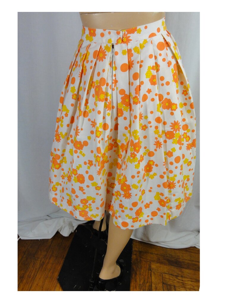 May include: A white skirt with a floral print in orange and yellow. The skirt has a pleated design and a zipper closure in the back.