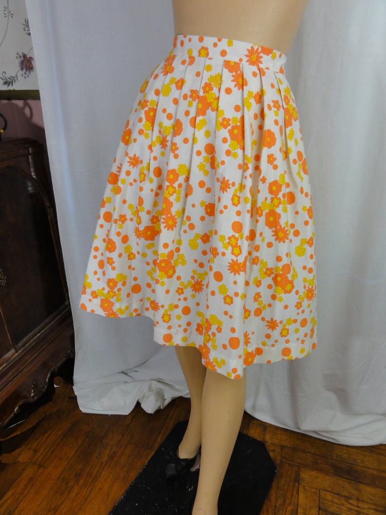May include: A white skirt with a pleated design and a floral print in orange and yellow. The skirt has a fitted waist and a flared hem.