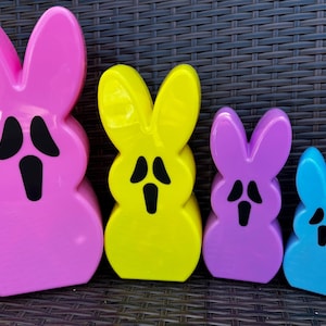 May include: Four brightly coloured bunny figurines in pink, yellow, purple, and blue. Each bunny has large ears and a black facial expression. The figurines are arranged in a row, decreasing in size.