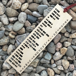 Puede incluir: Un marcapáginas de madera con un alfabeto braille y las letras N-Y-X-W-V-U-T-S-R-Q-P-O-N-M-L-K-J-I-H-G-F-E-D-C-B-A impresas en él. El marcapáginas tiene una cuerda roja atada.