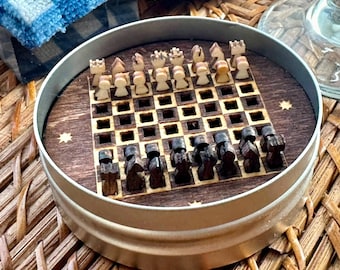 May include: A miniature chess set housed in a round metal tin. The board is made of dark wood with square cutouts, featuring light-colored wooden pieces on one side and dark-colored wooden pieces on the other. Small star-shaped inlays decorate the board.