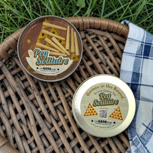 May include: Two round metal tins for a Peg Solitaire board game. One tin has a clear lid showing wooden pegs inside, labeled Peg Solitaire inGENEius. The other tin has a gold lid with the text Games in the Round Board Game Tin Peg Solitaire.