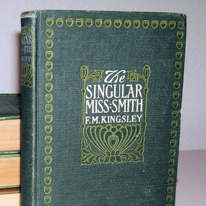 May include: A green hardcover book with gold lettering titled "The Singular Miss-Smith" by F.M. Kingsley. The book has a decorative border of gold apples.