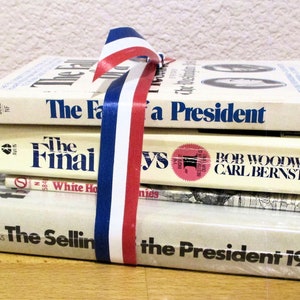 May include: A stack of four books about the presidency, wrapped in a red, white, and blue ribbon. The titles of the books are "The Fall of a President", "The Final Days", "The Selling of the President 1968", and "The Final Days".