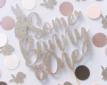 May include: Silver glitter cake topper with the text "Some bunny is one" and a small heart. The topper is surrounded by pink, brown, and white confetti circles and silver glitter bunny cutouts.