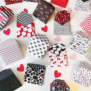 May include: A collection of small, folded paper envelopes with various patterns, including hearts, cherries, polka dots, arrows, and geometric designs. The envelopes are in shades of red, black, and white.