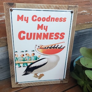 May include: A vintage metal sign with a wooden frame featuring a pelican with a large beak holding a row of Guinness bottles. The sign reads "My Goodness My Guinness".