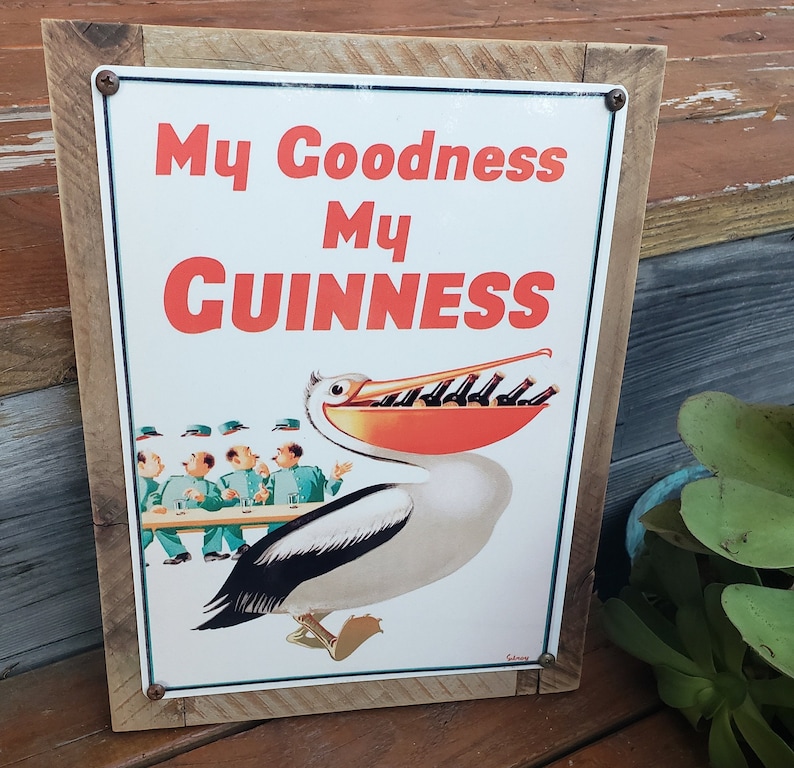 May include: A vintage metal sign with a wooden frame featuring a pelican with a large beak holding a row of Guinness bottles. The sign reads "My Goodness My Guinness".