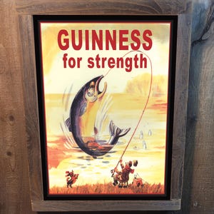 May include: A vintage-style metal sign with a brown wooden frame. The sign features a colorful illustration of a fisherman reeling in a large fish. The text on the sign reads "GUINNESS for strength".