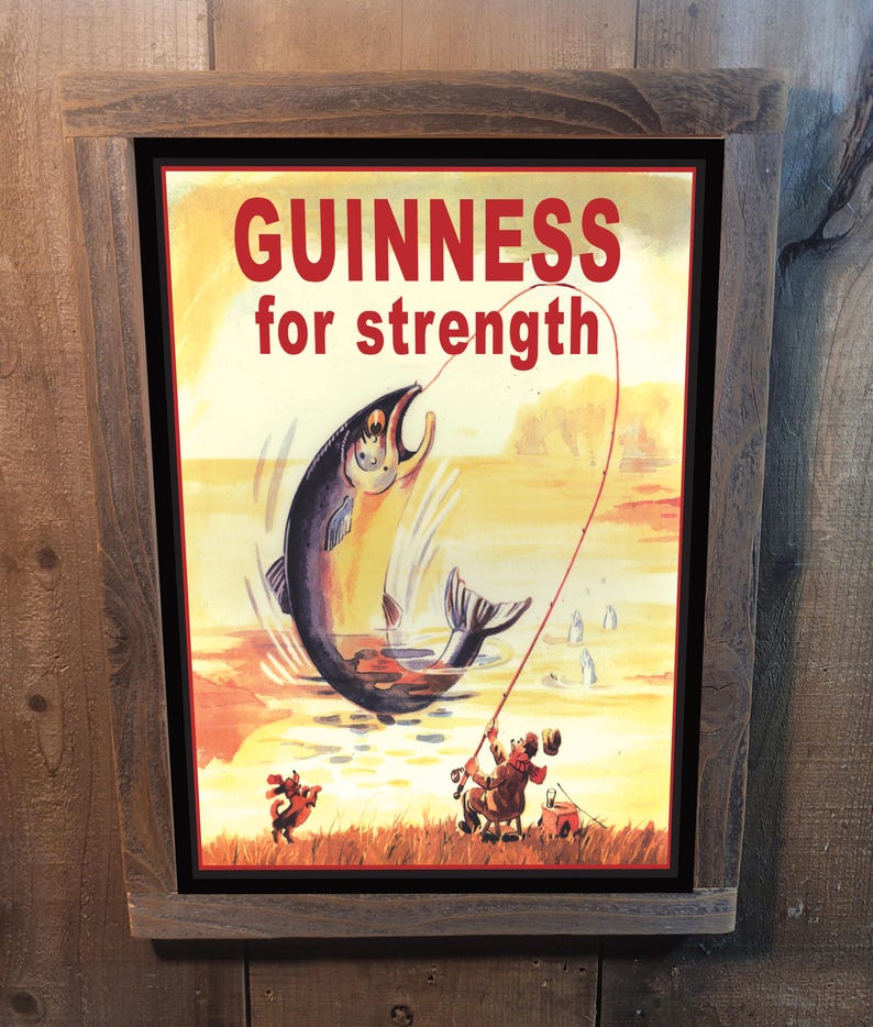 May include: A vintage-style metal sign with a brown wooden frame. The sign features a colorful illustration of a fisherman reeling in a large fish. The text on the sign reads "GUINNESS for strength".