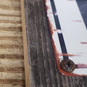 Puede incluir: Un marco de madera r&uacute;stico con un letrero blanco en el interior. El letrero tiene un borde negro y rojo y se ven dos tornillos.