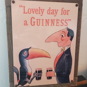 K&ouml;nnte beinhalten: Ein Vintage-Schild aus Metall mit einem Holzrahmen. Das Schild zeigt einen Cartoon-Tukan und einen Mann, der Gl&auml;ser Guinness h&auml;lt. Der Text auf dem Schild lautet "Lovely day for a GUINNESS".