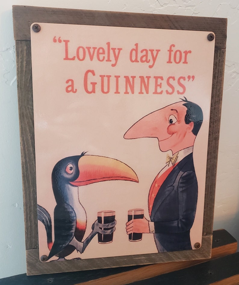 K&ouml;nnte beinhalten: Ein Vintage-Schild aus Metall mit einem Holzrahmen. Das Schild zeigt einen Cartoon-Tukan und einen Mann, der Gl&auml;ser Guinness h&auml;lt. Der Text auf dem Schild lautet "Lovely day for a GUINNESS".