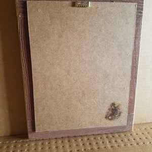 May include: Back of a framed artwork with a brown wooden frame. The back is made of a light brown cardboard material. A metal sawtooth hanger is attached to the top. The lower right corner has a stamp that says "Made in USA" and "Recycled Paper".