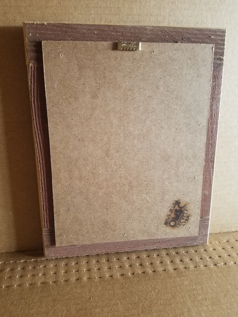 May include: Back of a framed artwork with a brown wooden frame. The back is made of a light brown cardboard material. A metal sawtooth hanger is attached to the top. The lower right corner has a stamp that says "Made in USA" and "Recycled Paper".
