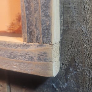 May include: Close-up of a corner of a rustic wooden picture frame. The frame is made of reclaimed wood with a natural gray finish. The wood has a rough texture and shows signs of wear and tear.