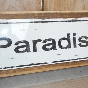 May include: A rustic wooden sign with a white metal plaque that reads "Paradise". The sign has a distressed look with a weathered brown finish.