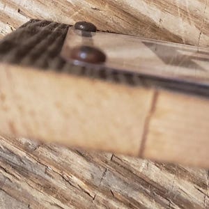 May include: Close-up of a wooden frame with a clear glass or plastic cover. Two small, dark-colored fasteners secure the cover to the frame. The wood has a natural, light brown color with visible grain.