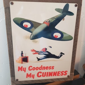 May include: A vintage metal sign with a green and white image of a plane flying over a man running towards a glass of beer. The sign reads "My Goodness My Guinness".