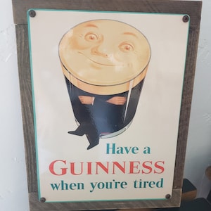 Puede incluir: Un letrero de metal vintage con marco de madera que anuncia la cerveza Guinness. El letrero presenta una ilustraci&oacute;n de dibujos animados de un vaso de Guinness con una cara sonriente y el texto "Have a GUINNESS when you're tired."