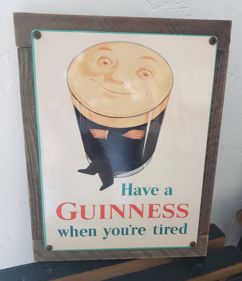 Puede incluir: Un letrero de metal vintage con marco de madera que anuncia la cerveza Guinness. El letrero presenta una ilustraci&oacute;n de dibujos animados de un vaso de Guinness con una cara sonriente y el texto "Have a GUINNESS when you're tired."