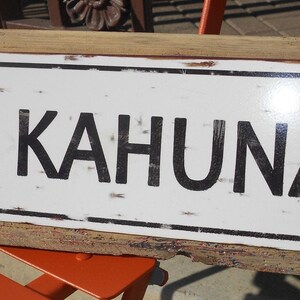 May include: A rustic wooden sign with a white metal plaque that reads "BIG KAHUNA". The sign has a distressed, weathered look with a rusty metal frame.
