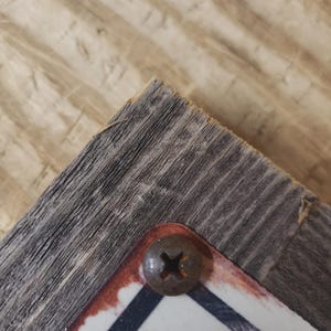 May include: Close-up of a corner of a weathered wooden sign with a rusty screw. The sign has a white background with a black and red border. The wood has a gray, textured appearance.