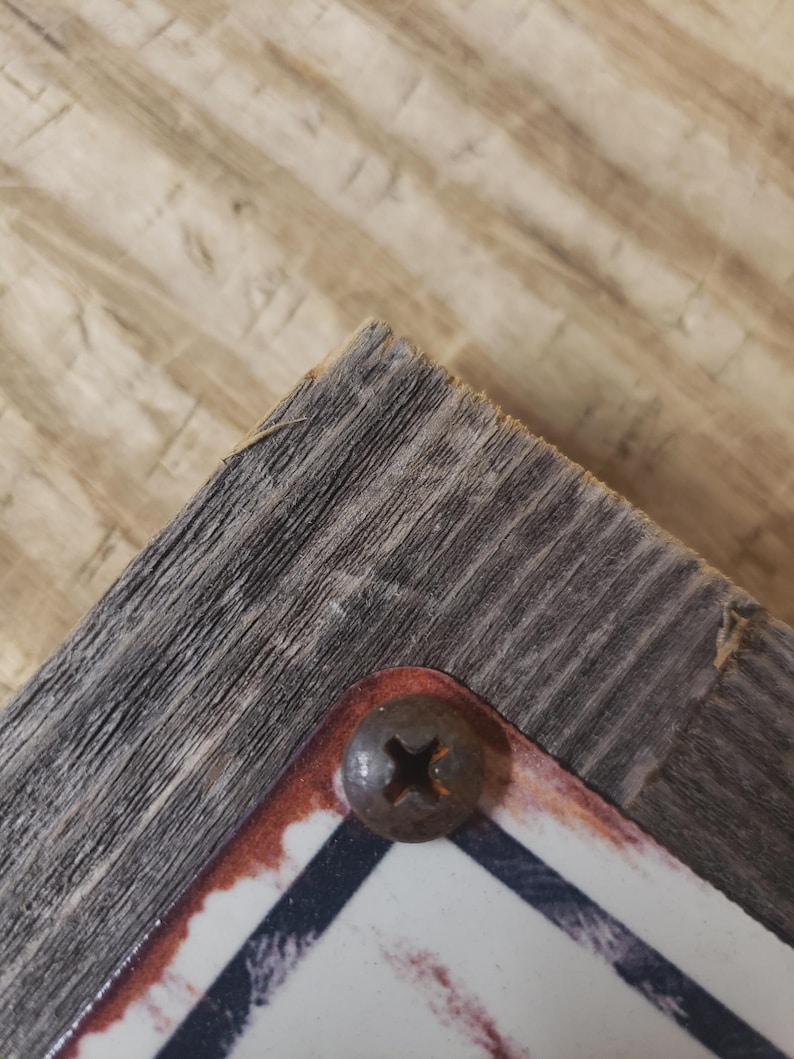 May include: Close-up of a corner of a weathered wooden sign with a rusty screw. The sign has a white background with a black and red border. The wood has a gray, textured appearance.
