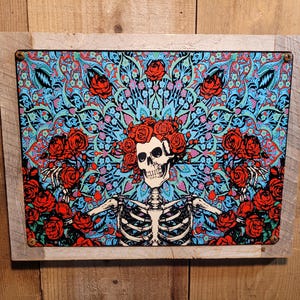 May include: A framed metal sign featuring a skeleton with a floral crown and rib cage, surrounded by red roses and a blue and teal patterned background. The sign is set in a rustic wooden frame.