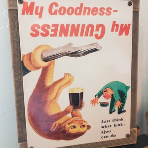 May include: A vintage metal sign with a brown wooden frame. The sign features a cartoon monkey holding a glass of Guinness beer. The text on the sign reads "My Goodness - my GUINNESS".  The sign also includes the text "Just think what kink - a jou can do".