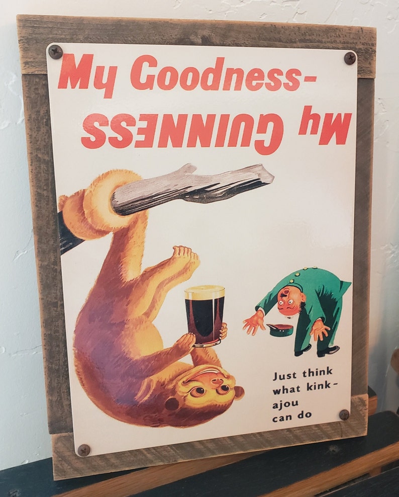 May include: A vintage metal sign with a brown wooden frame. The sign features a cartoon monkey holding a glass of Guinness beer. The text on the sign reads "My Goodness - my GUINNESS".  The sign also includes the text "Just think what kink - a jou can do".