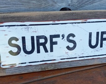 May include: A rustic wooden sign with a white metal plaque that reads "SURF'S UP" in black lettering. The sign has a distressed look with visible wood grain and rusty metal accents.