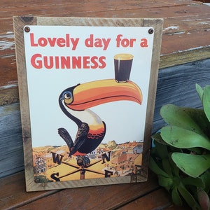 May include: A wooden sign with a brown background and a white rectangle in the center. The rectangle has a toucan with an orange beak holding a glass of Guinness. The text on the sign reads 