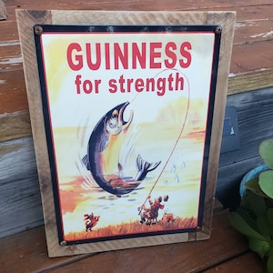 May include: A vintage metal sign with a wood frame advertising Guinness beer. The sign features a cartoon illustration of a large fish being caught on a fishing line. The text on the sign reads "GUINNESS for strength".