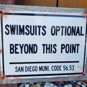 May include: A white sign with black text that reads "Swimsuits Optional Beyond This Point" and "San Diego Muni. Code 56.53". The sign is framed in wood with rusty metal accents.