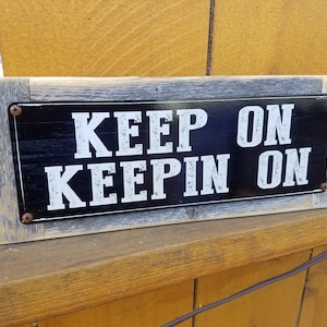 May include: A rustic wooden sign with a black metal plaque that reads "KEEP ON KEEPIN ON". The sign has a distressed wood finish and is secured with metal screws.
