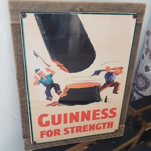 May include: A vintage metal sign with a wood frame featuring a cartoon illustration of two men chopping down a tree with the text "GUINNESS FOR STRENGTH".