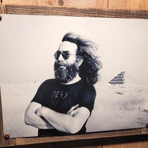 May include: Black and white framed print featuring a man with sunglasses and a beard, arms crossed. The man is wearing a t-shirt with Japanese characters. A pyramid is in the background. The frame is made of wood.